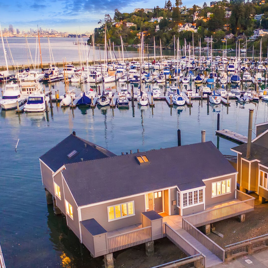 Waterfront home beside a marina, with sailboats behind it and warm light glowing through the windows.